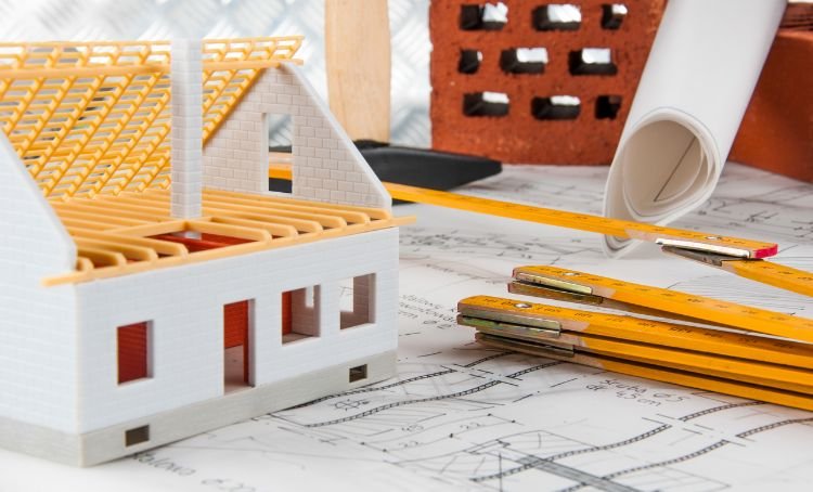 Blueprints new build property with a small toy house and pencils on it - Financing a New Home Build - ECF