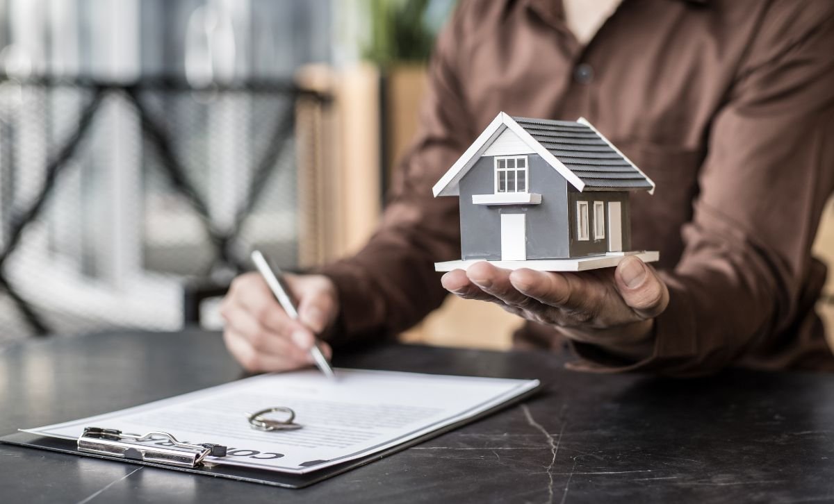 A hand holding a small model house while signing a contract - Express Capital Financing