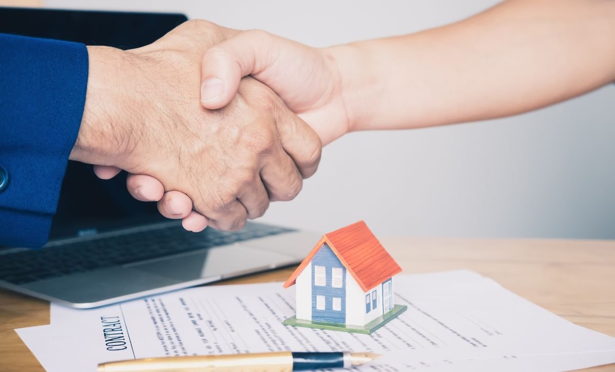 A close-up of a handshake over a desk with a contract and a small model of a house sitting on it - Express Capital Financing