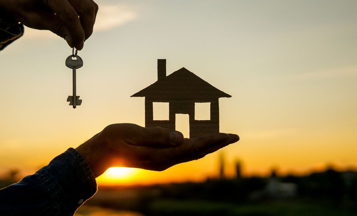A hand holding a small wooden house with a key in the other hand, in front of a sunset - Express Capital Financing